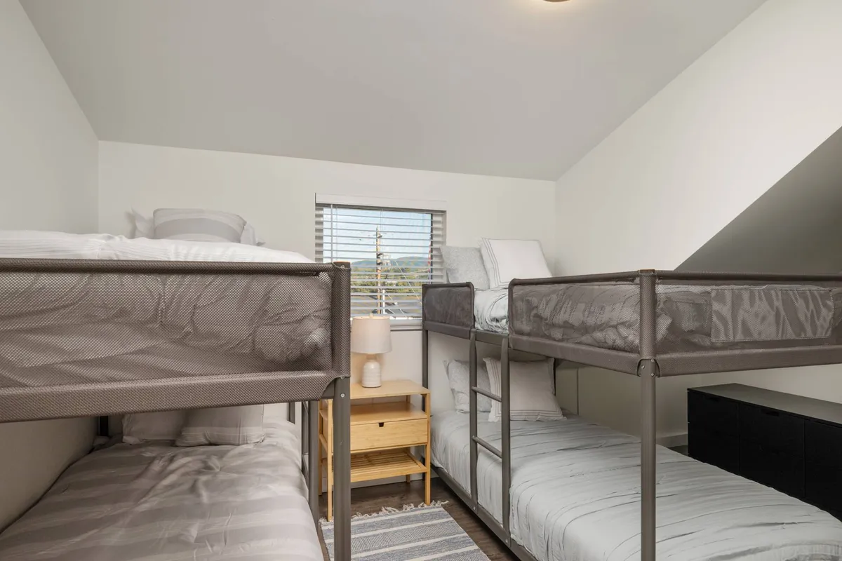 A cheerful bunk room with two sets of twin-over-twin bunk beds, a window that lets in plenty of daylight, a nightstand, and a lamp. The arrangement invites late-night chats and creates a playful, social atmosphere ideal for kids or groups of friends.