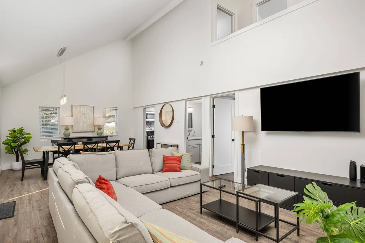 A cozy and modern open-concept living and dining area featuring a comfortable sectional sofa, a large wooden dining table, a big TV, a decorative plant, and ample light. The layout encourages togetherness and flows seamlessly between spaces, with plush furnishings and tasteful decor that evoke a coastal vibe.