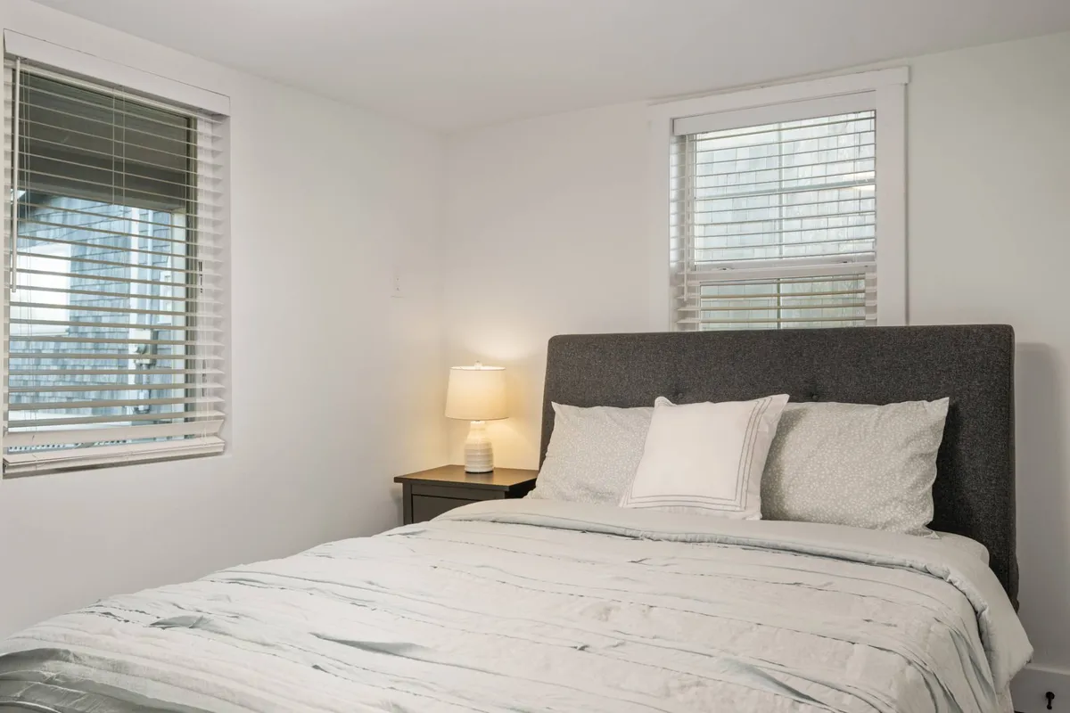A serene bedroom featuring a queen-size bed with plush, soft linens, a bedside lamp on a nightstand, and windows that let in abundant natural light. The color palette is soothing, with modern touches and comfortable furnishings aimed at relaxation.