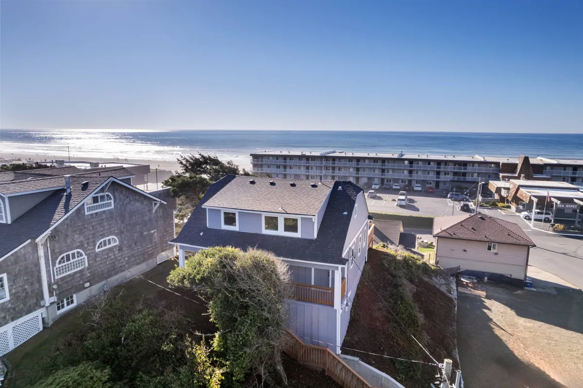 The image showcases the remodeled exterior of a stylish single-family house perched above the Lincoln City coastline, with sweeping ocean views, a spacious new deck, and the serene natural landscape of the Oregon coast. The deck is invitingly set for gatherings and relaxation, while the home's facade hints at modern upgrades and comfort.