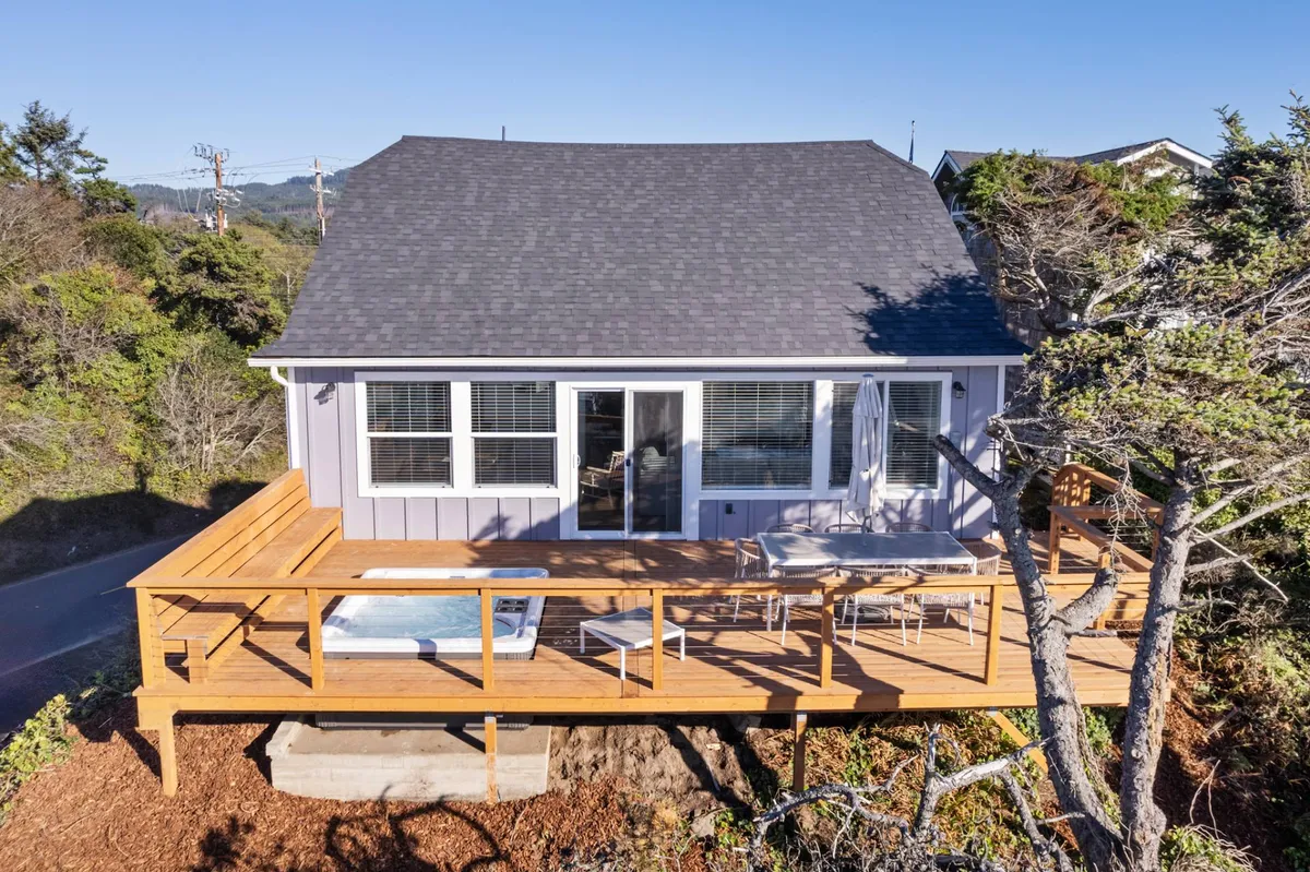 This inviting exterior image showcases the large wooden deck, complete with a hot tub and surrounded by lush trees. The deck is a focal point for relaxation, outdoor dining, and communal gatherings, all set against a backdrop of tranquil nature.