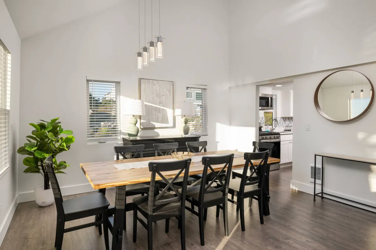 A warm, inviting dining area with a large wooden table set beneath stylish light fixtures. Decorative elements like wall art, a mirror, and a potted plant add personality while oversized windows bathe the space in natural light, enhancing the coastal ambiance.