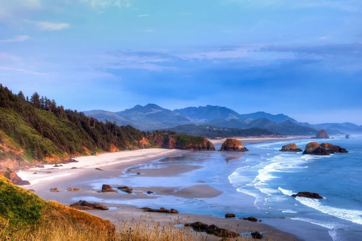 A stunning view of the Oregon coast, showcasing crashing ocean waves, dramatic rock formations, distant mountains, and a sandy beach. The image captures the natural beauty and outdoor appeal of Lincoln City, inviting exploration beyond the property itself.