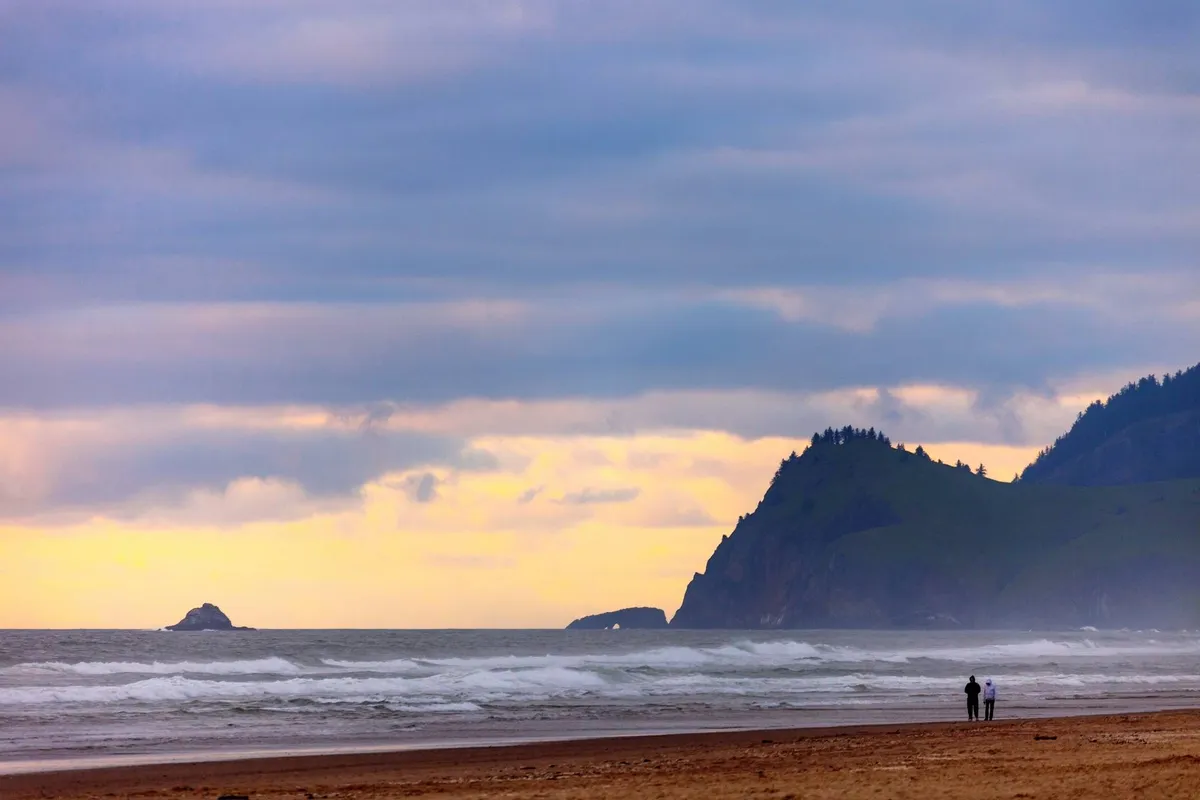 A sweeping beach view with distant cliffs, crashing waves, and a couple walking along the shoreline. The scene captures the natural beauty, opportunities for beachcombing, and room for family fun on the sand.
