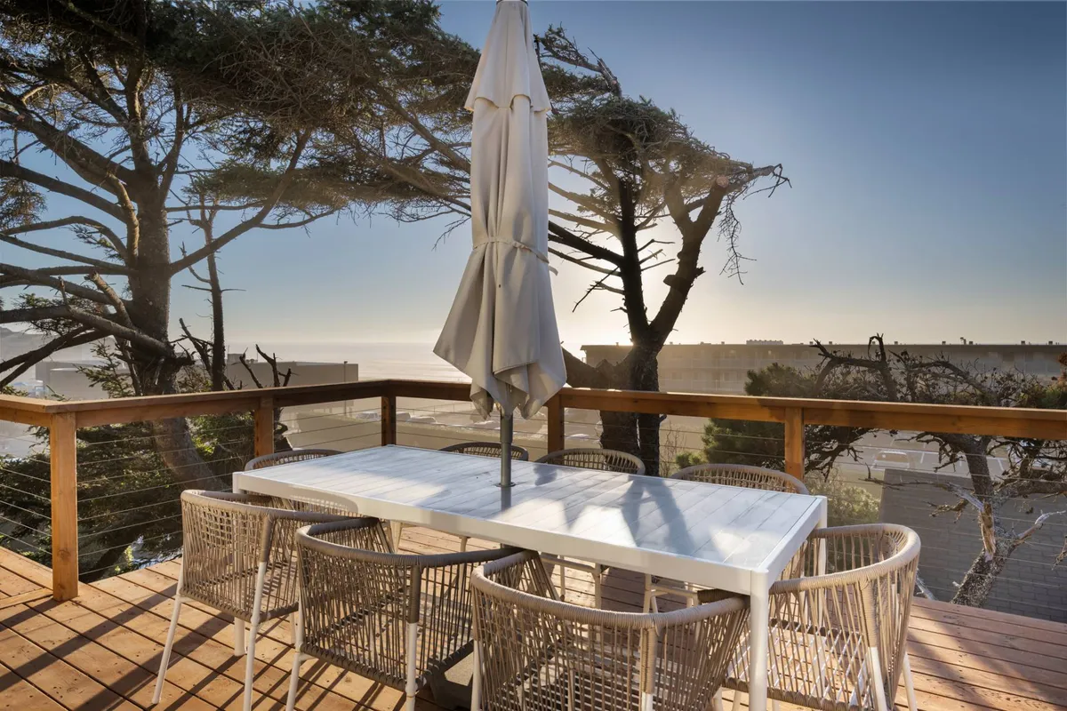 An exterior shot of a spacious wooden deck set against a backdrop of trees and ocean views. The focal point is an outdoor dining table with an umbrella, ideal for al fresco meals, relaxing, or gathering with friends and family.
