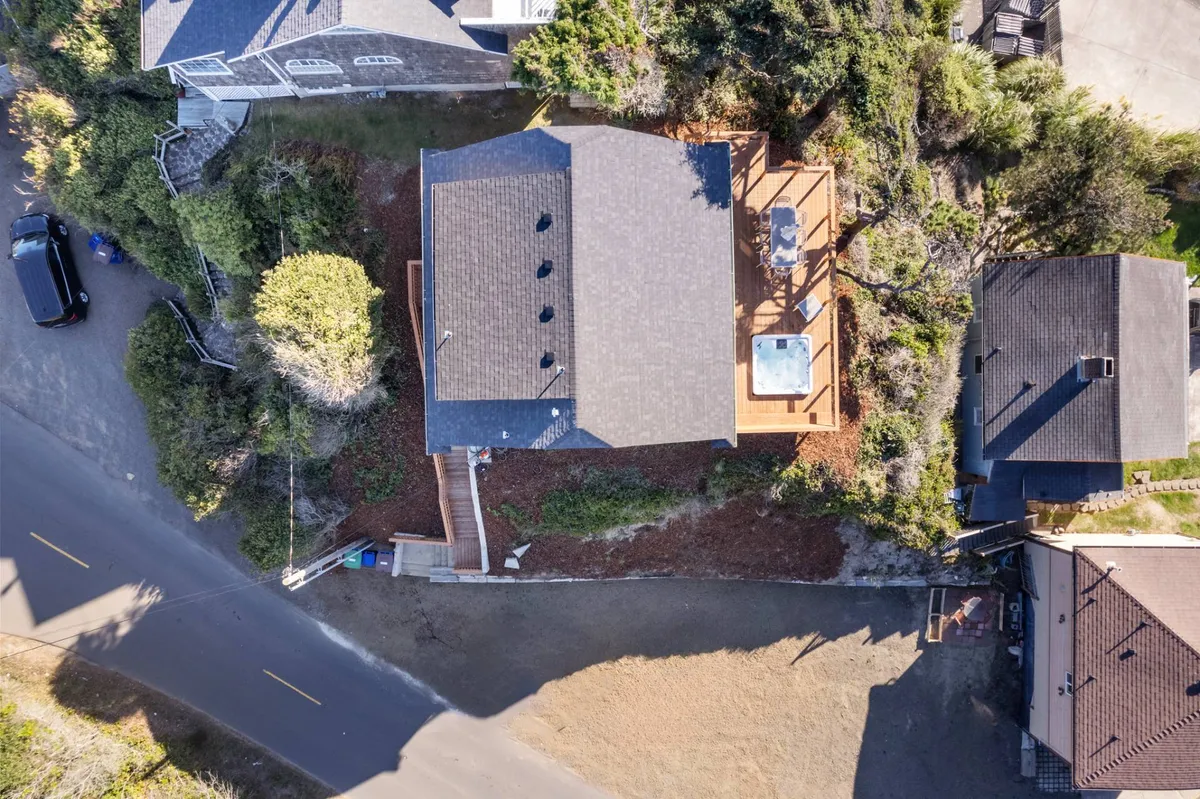 An exterior shot of the house structure featuring the deck area with a hot tub, surrounded by greenery and an adjacent road leading toward nature. The space looks welcoming for outdoor dining, relaxing, and as a jumping-off point for nearby walks or trips to local pet-friendly spots.