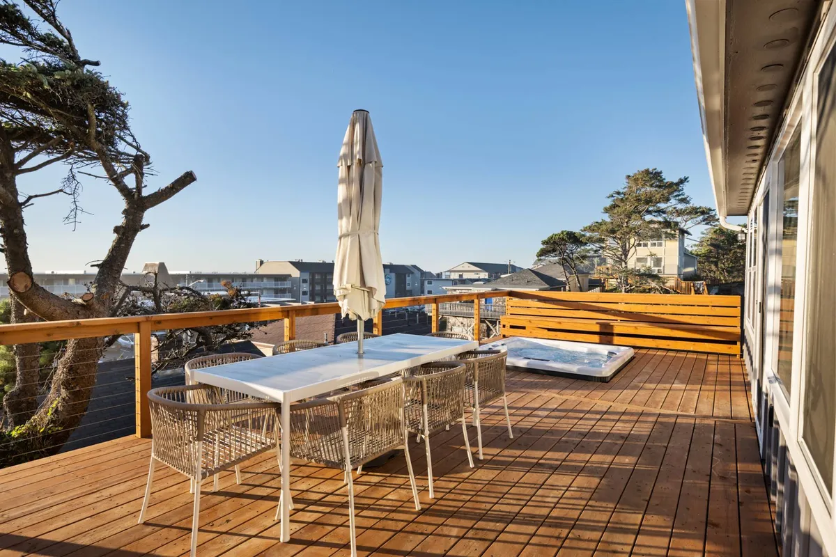 The image features a relaxed outdoor dining area on the wooden deck with a dining table, umbrellas, and a sweeping view of the neighborhood. The setup invites guests to enjoy fresh air meals and sociable moments outdoors.