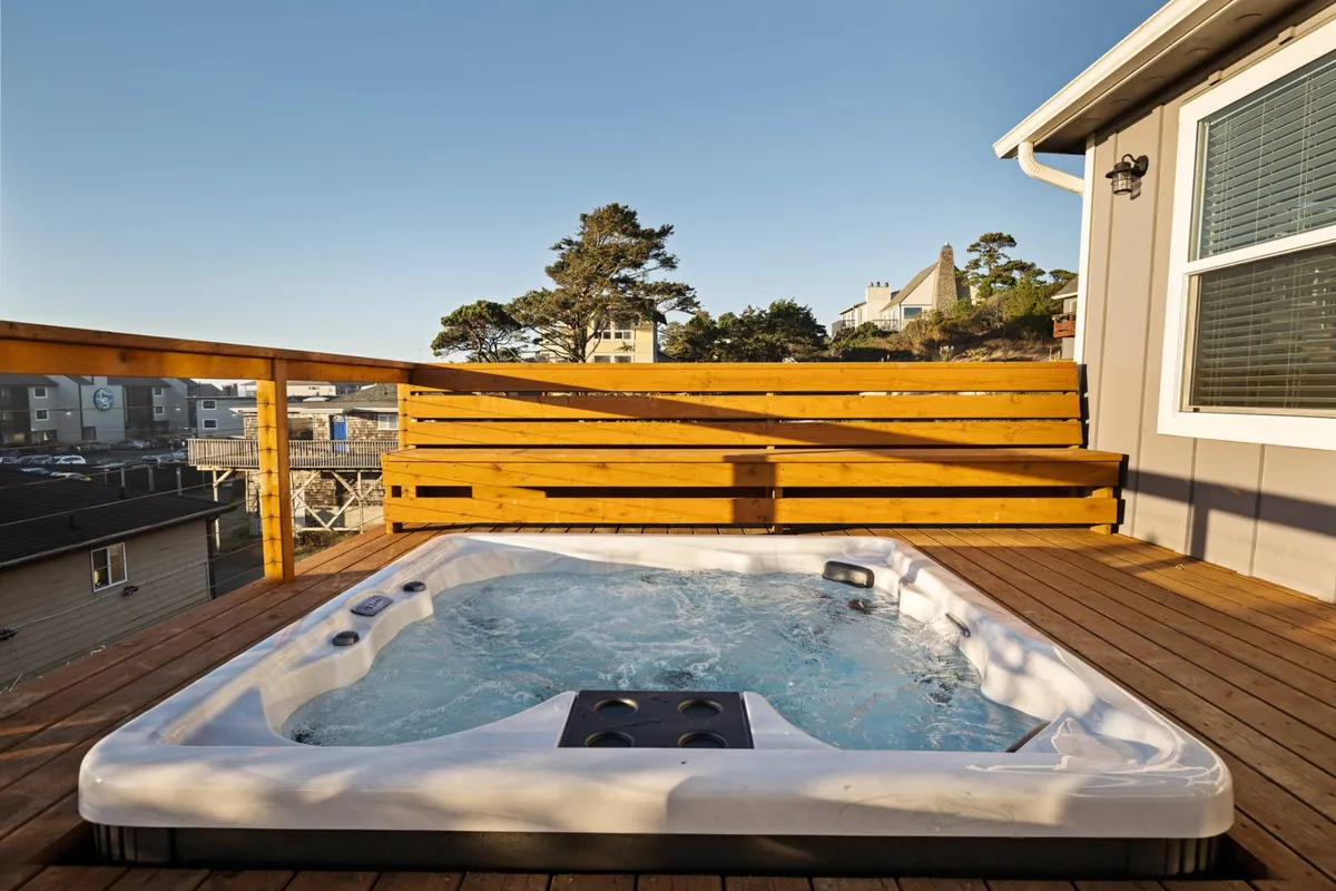 This image features a tranquil outdoor hot tub set on a wooden deck, surrounded by lush trees. The setting is ideal for relaxation, with enough space for a small group to unwind while taking in the natural views beyond the deck.