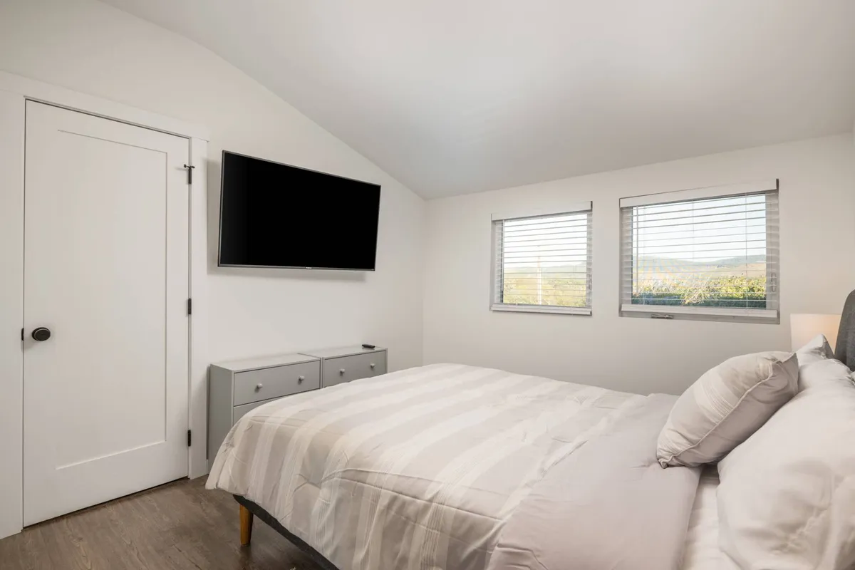 This inviting bedroom interior features a comfortable bed facing windows that let in natural light, a TV, and a dresser. The room suggests peaceful mornings with coffee, quiet evenings relaxing in bed, and a tranquil ambiance perfect for couples seeking a romantic retreat.